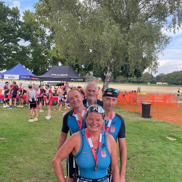 Gruppe von vier Triathleten mit Medaillen, nach einem Wettkampf im Freien, umgeben von Zuschauern und Pavillons.