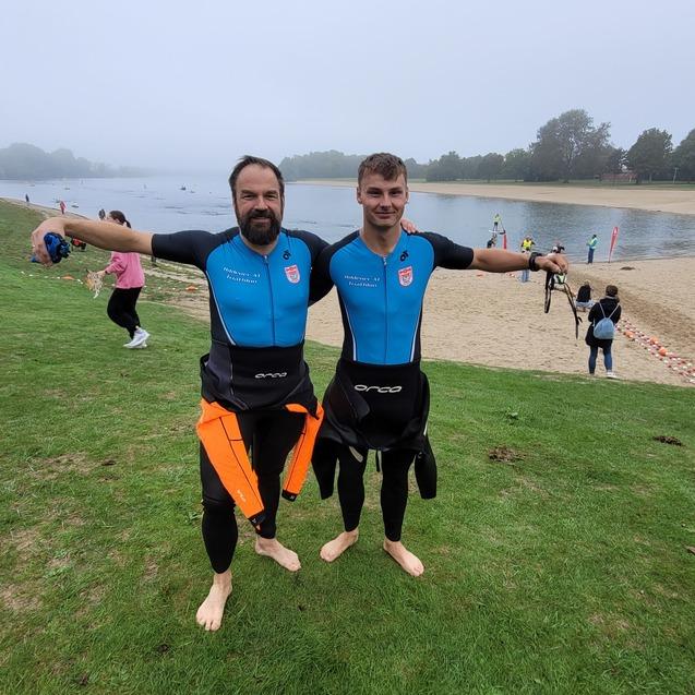Zwei Männer in Neoprenanzügen stehen mit ausgebreiteten Armen am Ufer eines Badestrandes, im Hintergrund sind Menschen im Wasser.