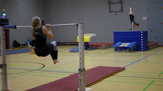 Zwei Turnerinnen im Training: Eine macht Klimmz&uuml;ge an den Stangen, die andere &uuml;bt einen Sprung vom Block.