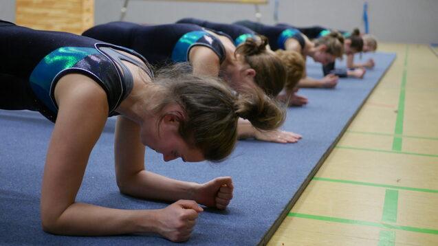 Eine Gruppe von Turnerinnen macht Planks auf einer blauen Trainingsmatte in einer Sporthalle.