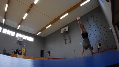Gymnastin f&uuml;hrt einen Handstand auf einem blauen Turnfeld in einer Sporthalle, im Hintergrund sind Zuschauer und eine Kletterwand.