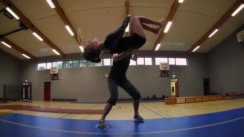 Ein T&auml;nzer hebt eine Partnerin in einer Sporthalle, umgeben von Turnger&auml;ten und Fenstern.