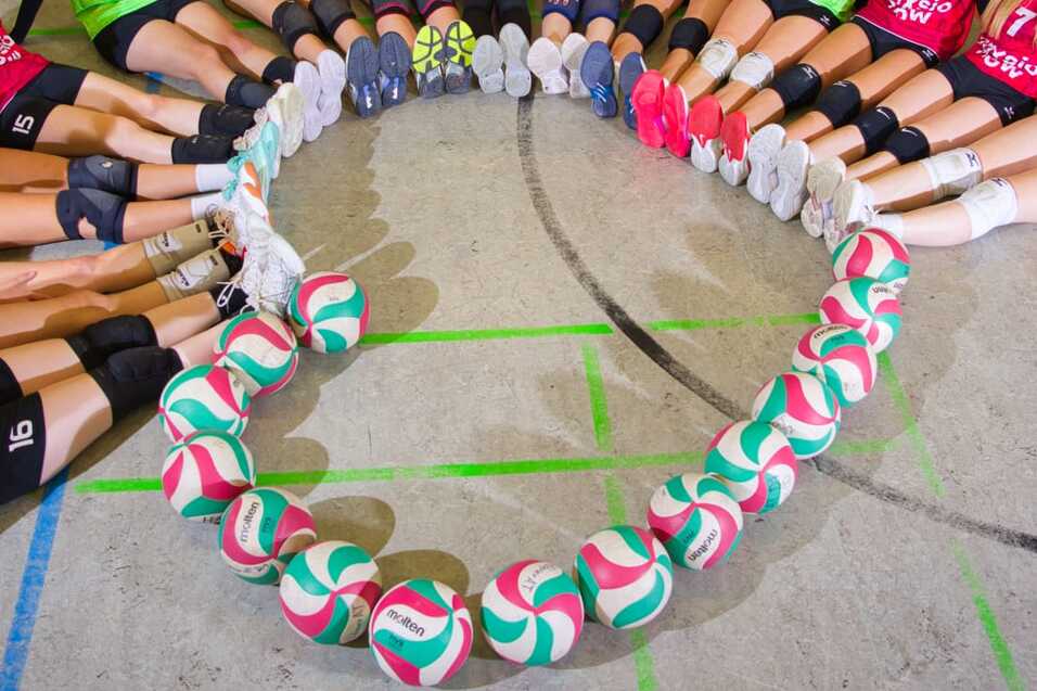 Gruppe von Volleyballspielerinnen in roten Trikots sitzt um einen Kreis aus bunten Bällen in einer Sporthalle.