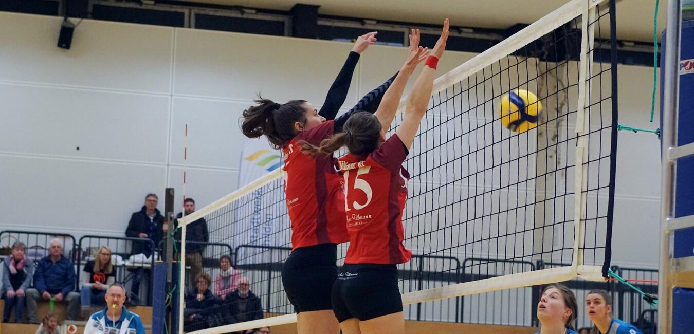 Zwei Volleyballspielerinnen blockieren einen Ball &uuml;ber dem Netz in einer Sporthalle w&auml;hrend eines Spiels.