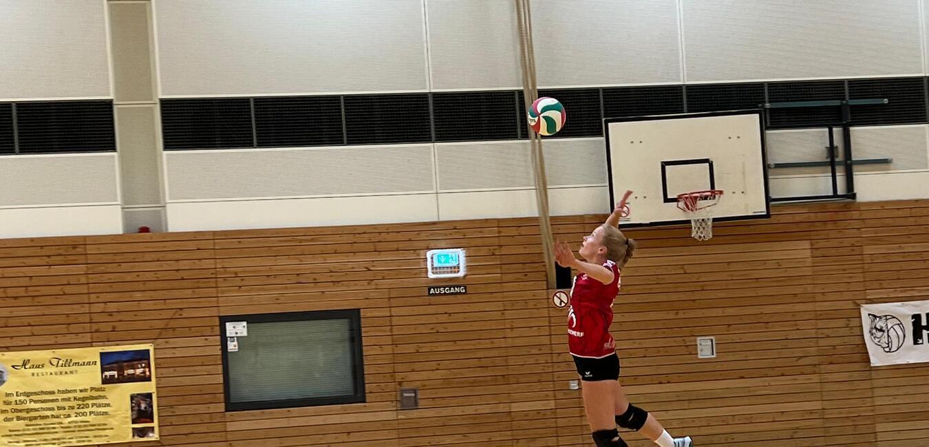 Volleyballspieler in rotem Trikot springt hoch, um einen Ball &uuml;ber das Netz in einer Sporthalle zu schlagen.