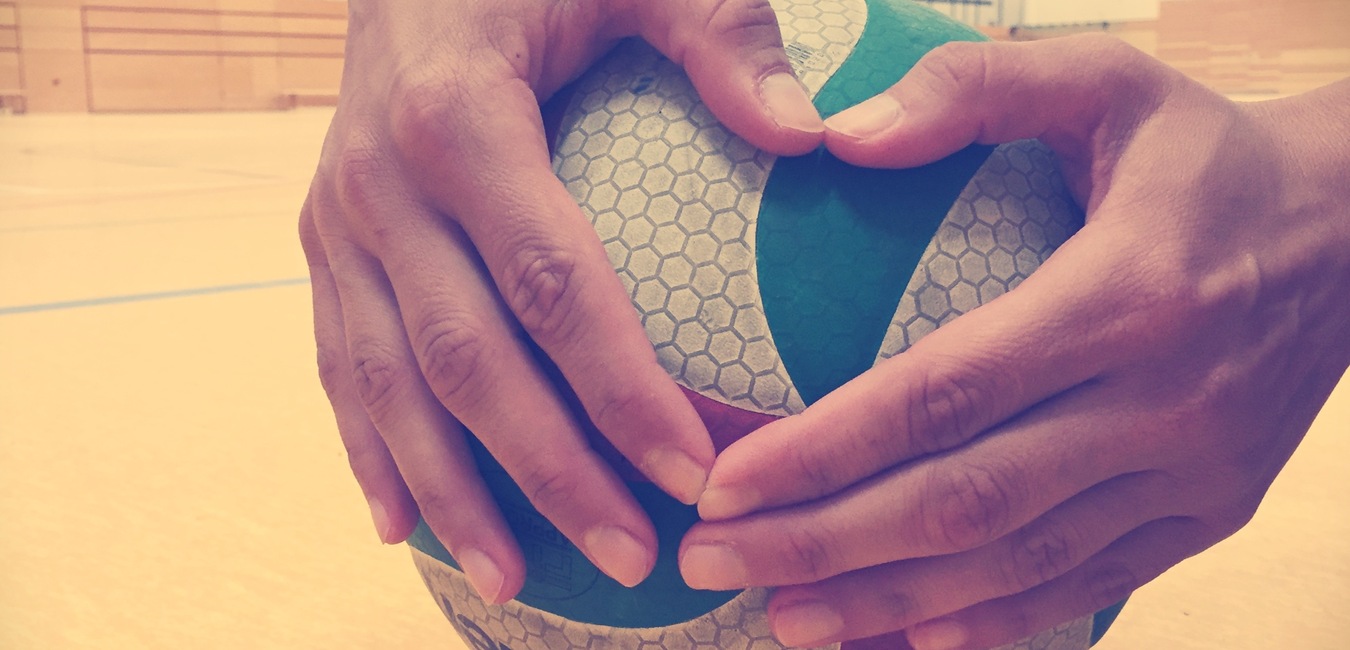 H&auml;nde halten einen Volleyball, der auf einem Sportboden liegt, im Hintergrund sind Sporteinrichtungen sichtbar.