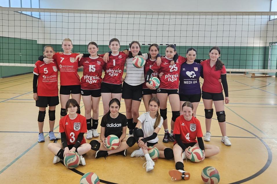 Gruppierungsfoto eines Volleyballteams mit 15 M&auml;dchen in rot-schwarzen Trikots, umgeben von Volleyballen auf dem Spielfeld.