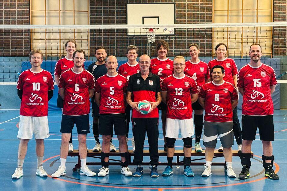 Gruppenaufnahme von zwölf Volleyballspielern in roten Trikots in einer Sporthalle, mit Netz im Hintergrund.