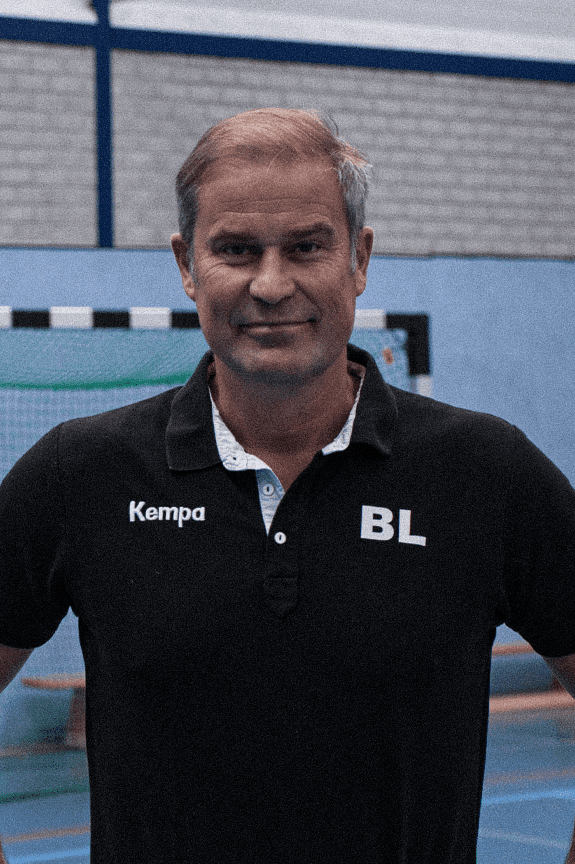Mann in sportlicher Kleidung steht in einer Sporthalle, hinter ihm ein Handballtor. Logo und Text sind im Bild integriert.