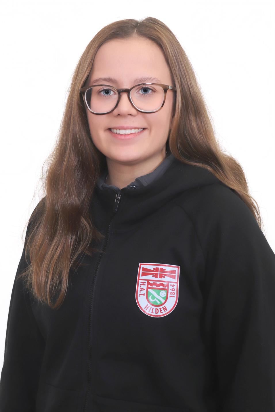 Porträt einer lächelnden jungen Frau mit langen, lockigen Haaren, die einen schwarzen Hoodie mit Logo trägt.