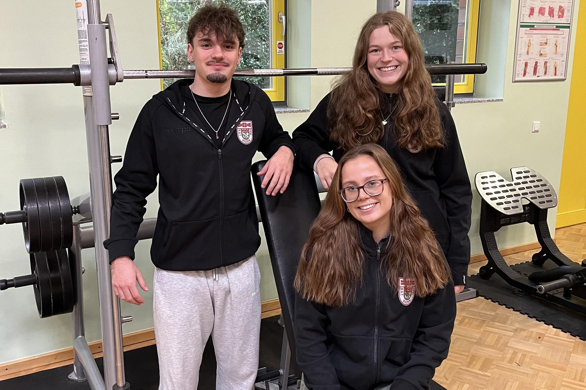 Drei junge Menschen in Sportkleidung stehen in einem Fitnessraum vor einem Kraftger&auml;t.
