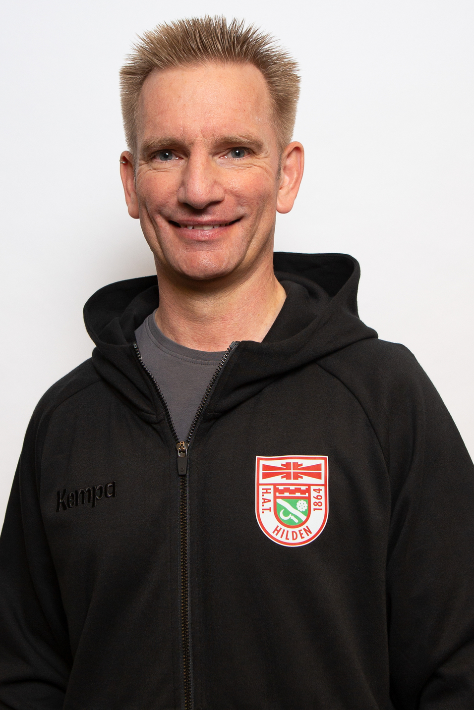 Mann mit kurzem, blondem Haar in einem schwarzen Hoodie mit Logo steht vor einem wei&szlig;en Hintergrund und l&auml;chelt.