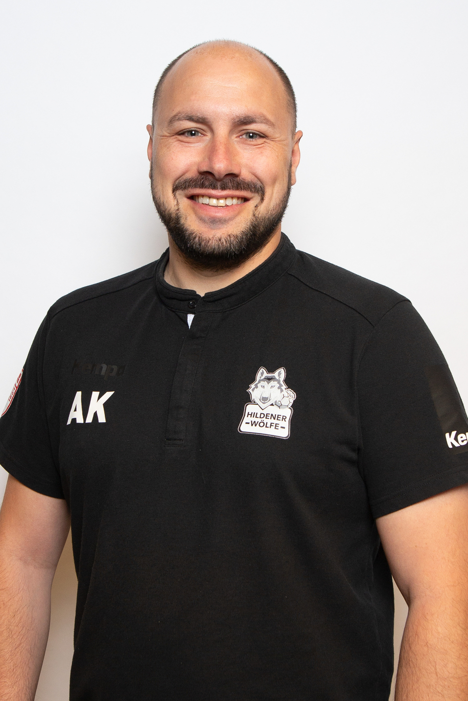Portr&auml;t eines l&auml;chelnden Mannes in schwarzem Sportshirt mit Logo und Initialen, vor einem wei&szlig;en Hintergrund.