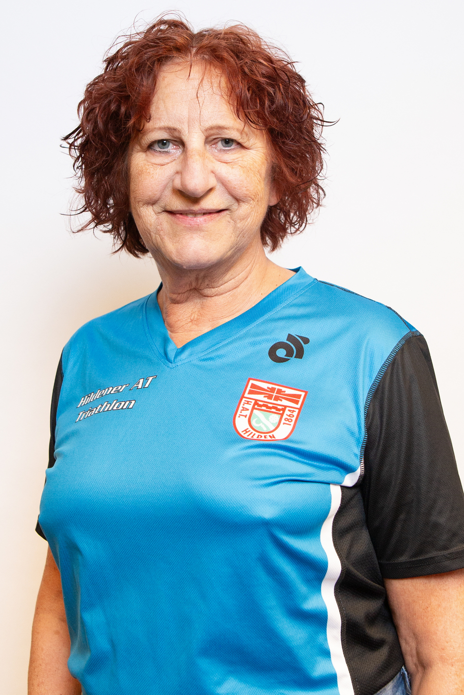 Portr&auml;t einer l&auml;chelnden Frau mit lockigem, rotem Haar, tr&auml;gt ein blaues Sportshirt mit Vereinslogo.