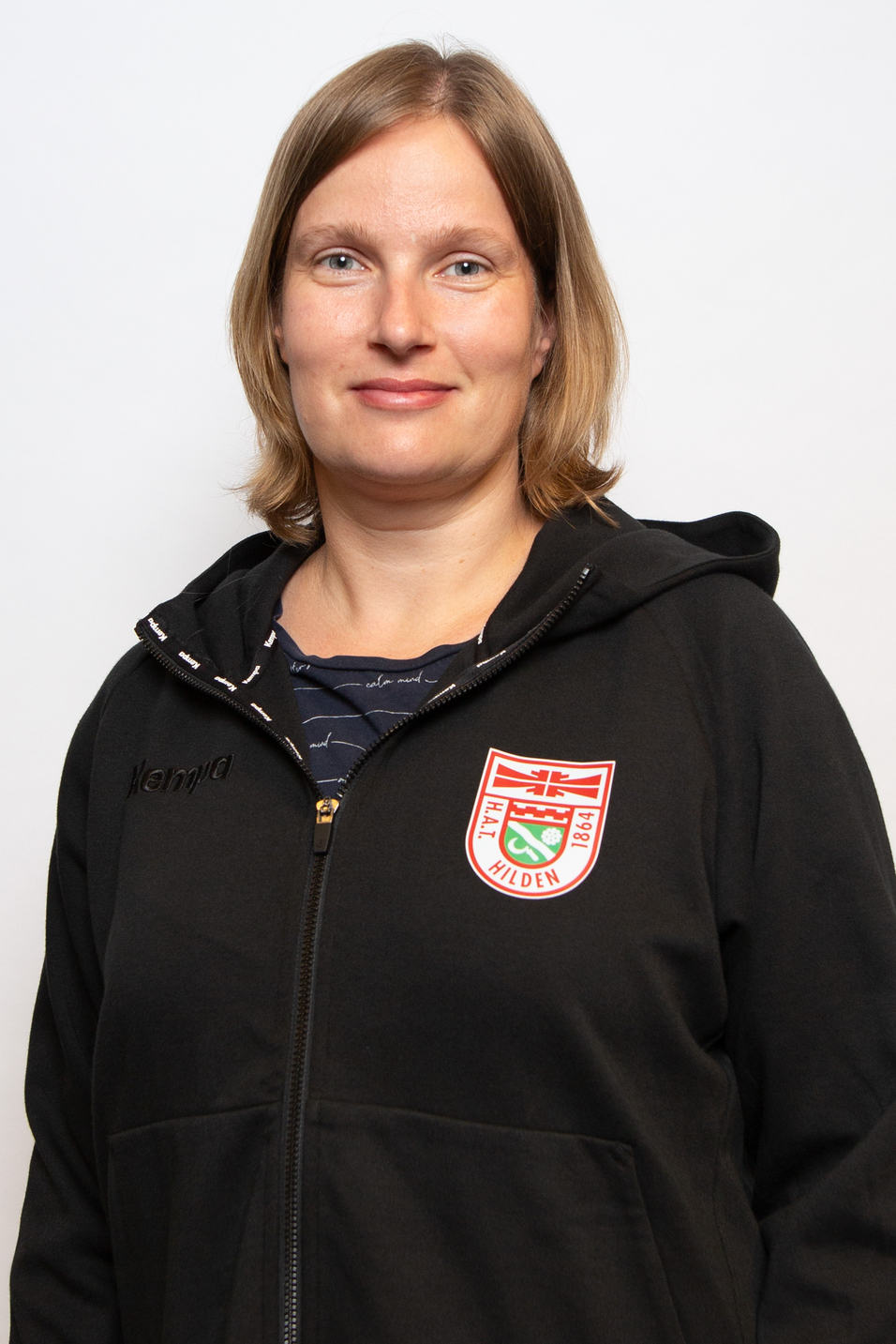 Portr&auml;t einer Frau mit blonden Haaren in einem schwarzen Kapuzenpullover mit Logo des Vereins &bdquo;Hallen Hilden&ldquo;.