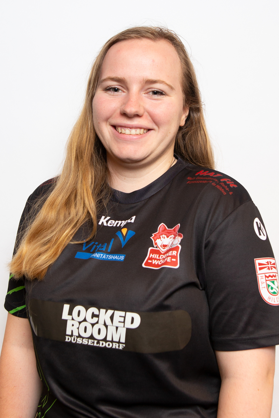 Portr&auml;t einer l&auml;chelnden Frau in einem schwarzen Sporttrikot mit Logos, vor einem neutralen Hintergrund.