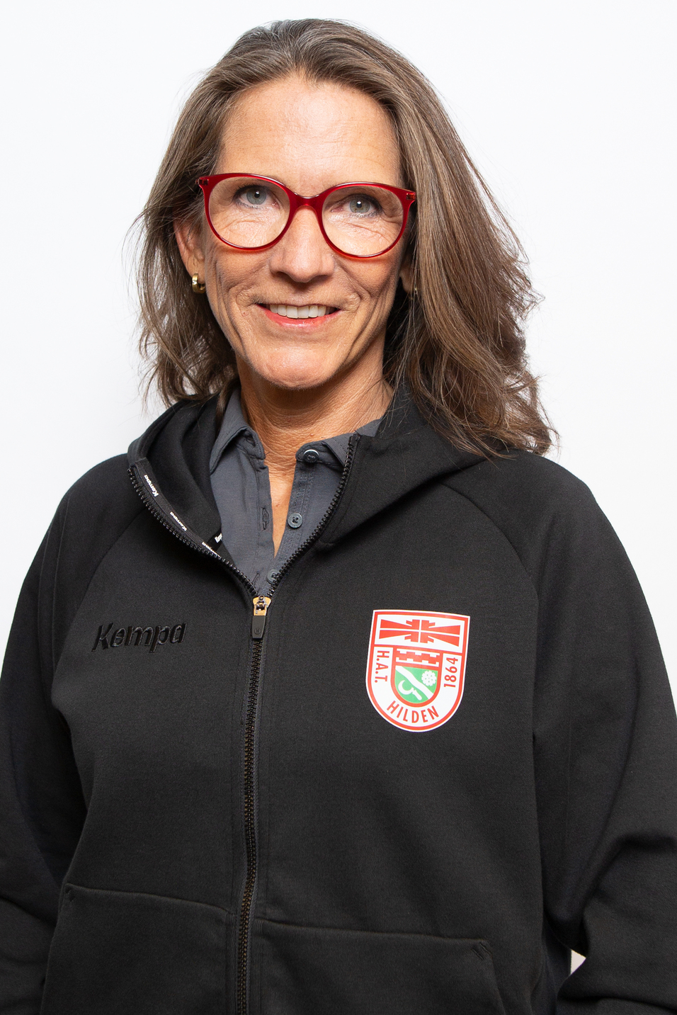 Portr&auml;t einer l&auml;chelnden Frau mit langen braunen Haaren, tragt eine schwarze Jacke mit Logo und rote Brille.