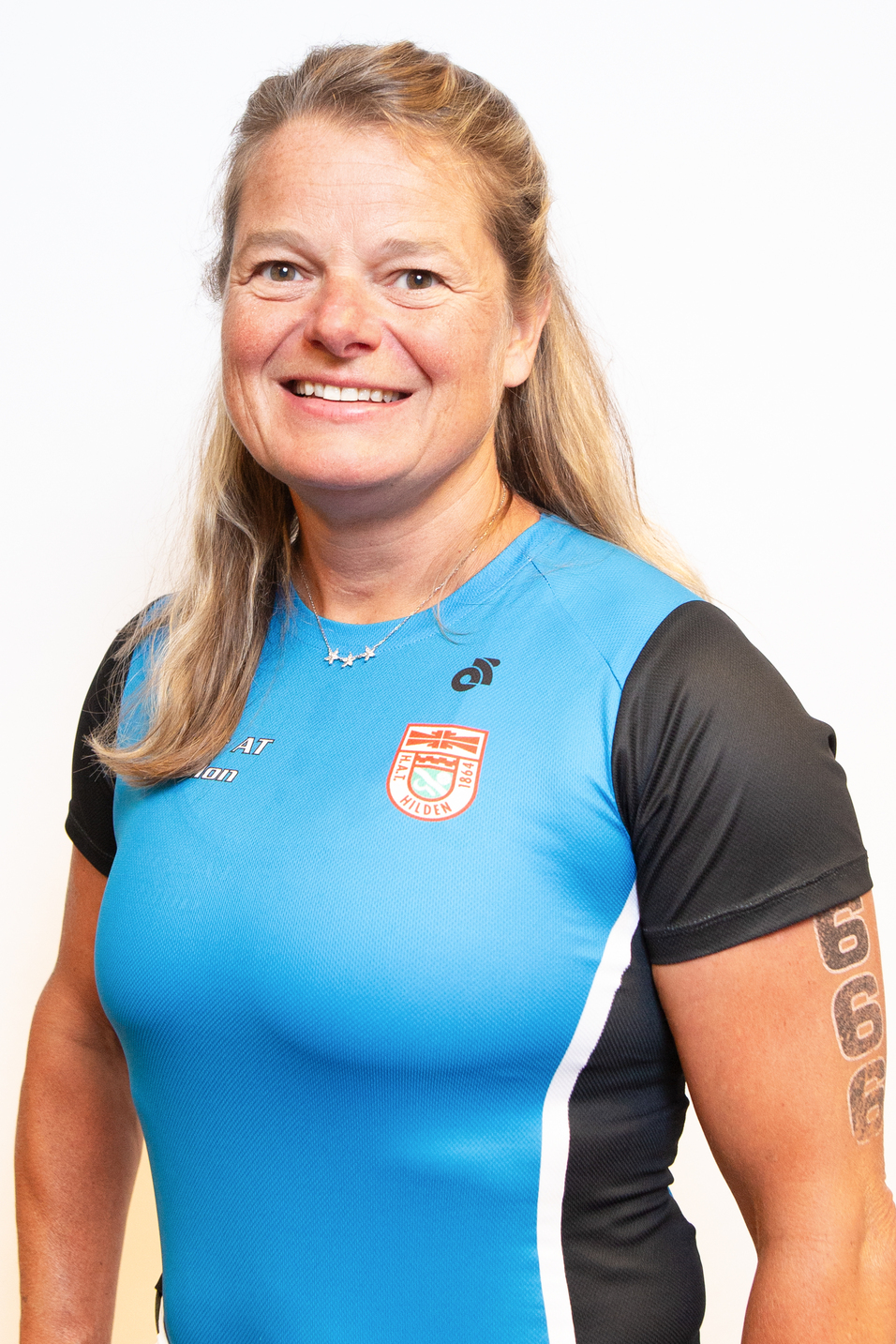 Portr&auml;t einer l&auml;chelnden Frau in einem blauen Sportshirt mit kurzem &Auml;rmel und T&auml;towierung am Arm vor einem wei&szlig;en Hintergrund.