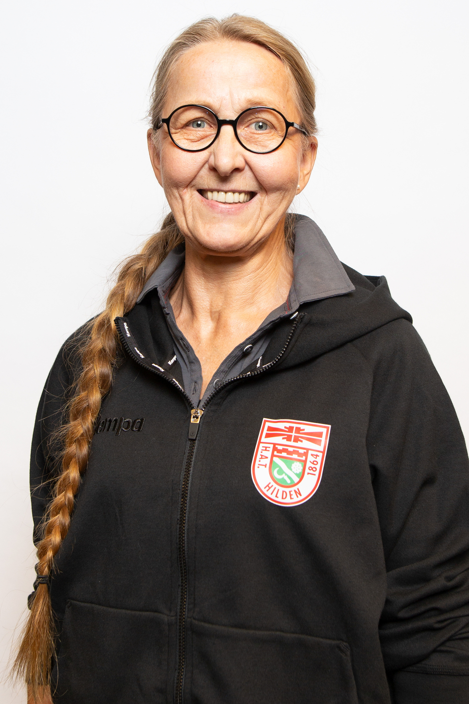 Portr&auml;t einer l&auml;chelnden Frau mit langen, geflochtenen Haaren, Brille und schwarzer Jacke mit Logo.