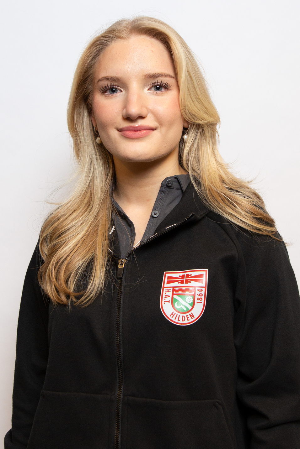 Portr&auml;t einer jungen Frau mit langen, blonden Haaren, die einen schwarzen Hoodie mit Logo tr&auml;gt und l&auml;chelt.
