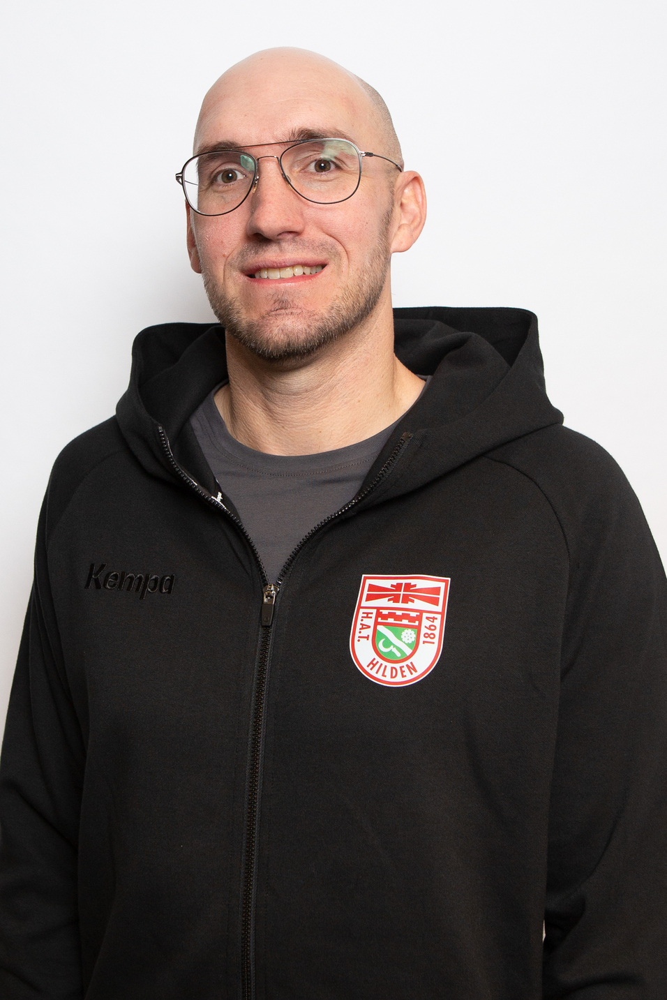 Portrait eines Mannes mit Glatze in schwarzer Kapuzenjacke, der l&auml;chelnd vor einem wei&szlig;en Hintergrund steht.