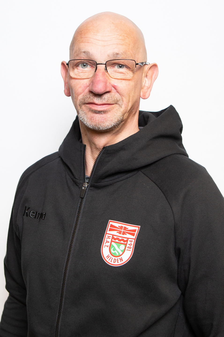 Portrait eines Mannes in schwarzer Kapuzenjacke mit Brille, l&auml;chelnd vor wei&szlig;em Hintergrund.