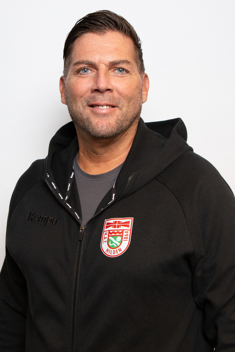 Mann mit kurzer Frisur und Bart l&auml;chelt in schwarzem Kapuzenpulli mit Logo vor neutralem Hintergrund.