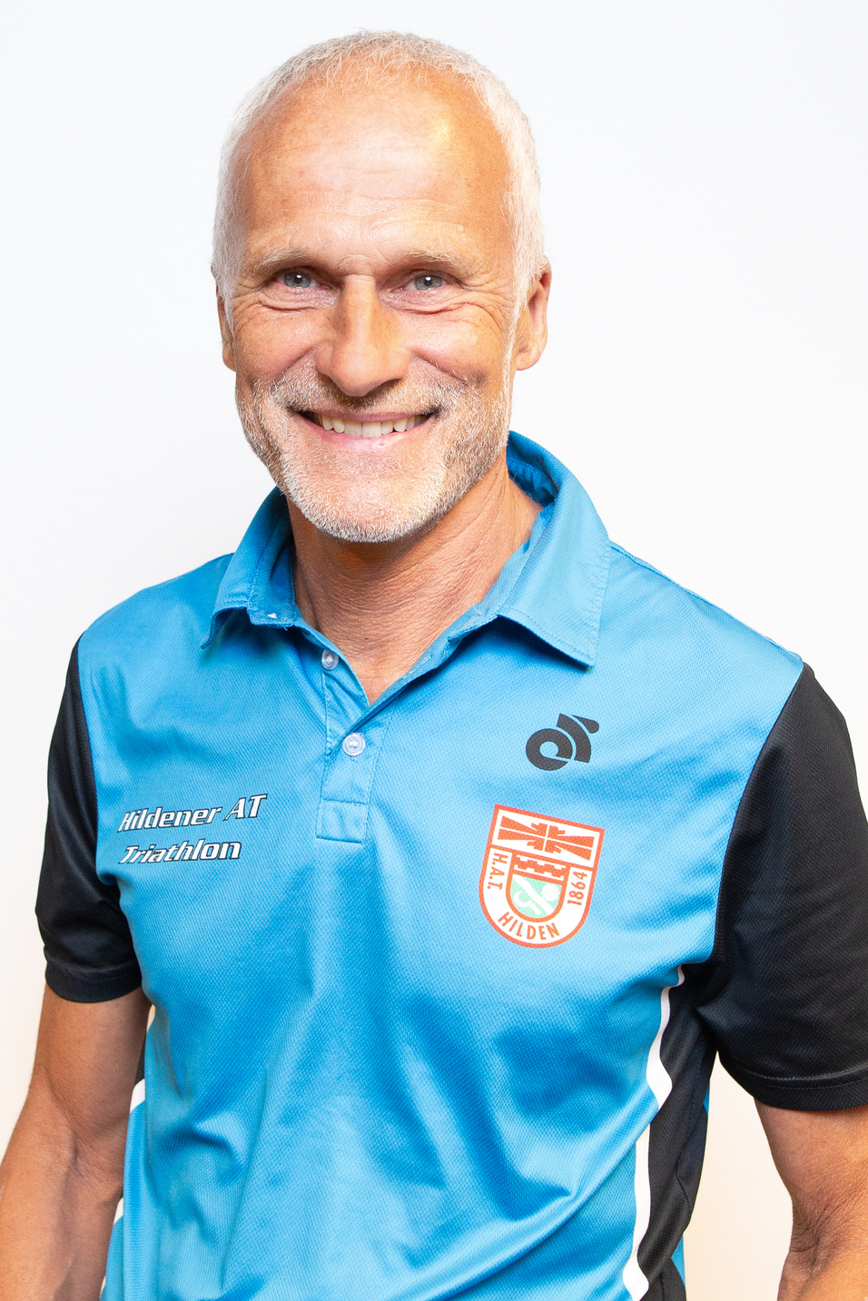 L&auml;chelnder Mann mit grauen Haaren tr&auml;gt ein blau-schwarzes Polo-Shirt mit Logo und Aufschrift &bdquo;Triathlon Hilden&ldquo;.