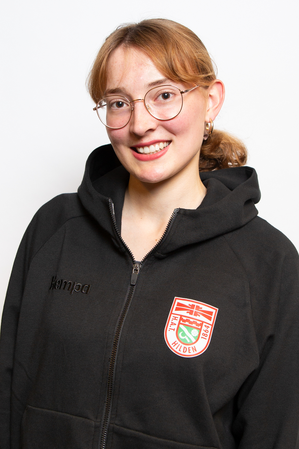 Junge Frau mit Brille und Zopf l&auml;chelt in einem schwarzen Hoodie mit Vereinswappen.