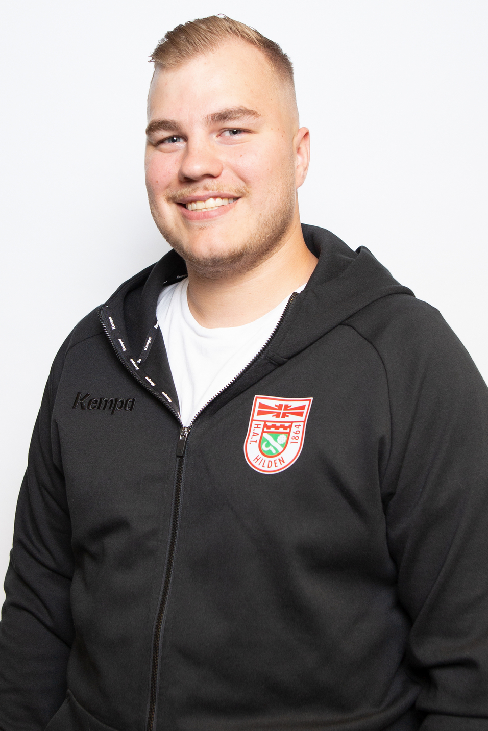 Junger Mann mit kurzem Haar und freundlichem L&auml;cheln, tr&auml;gt einen schwarzen Kapuzenpullover mit Vereinslogo.