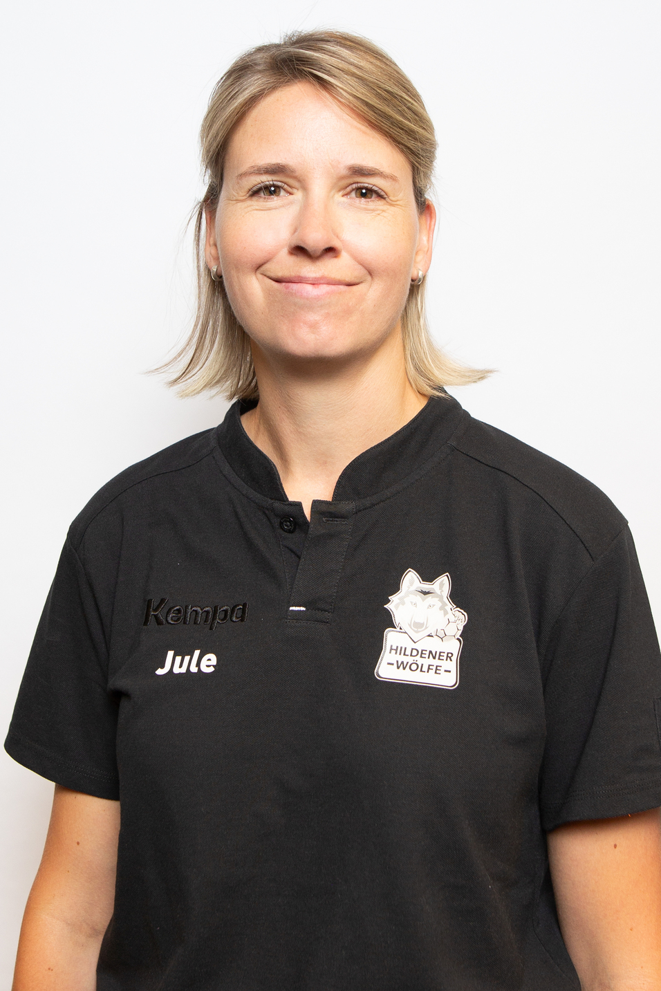 Portr&auml;t einer l&auml;chelnden Frau mit kurzem, blondem Haar, in schwarzem Polo-Shirt mit Aufdruck und Namensschild.