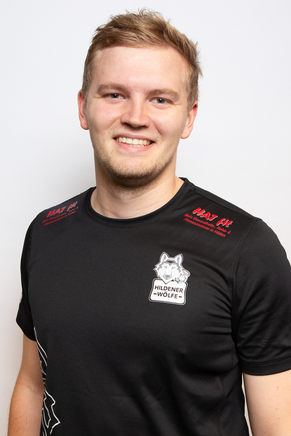 Portr&auml;t eines l&auml;chelnden Mannes in einem schwarzen T-Shirt mit dem Logo der Hildener W&ouml;lfe auf hellem Hintergrund.