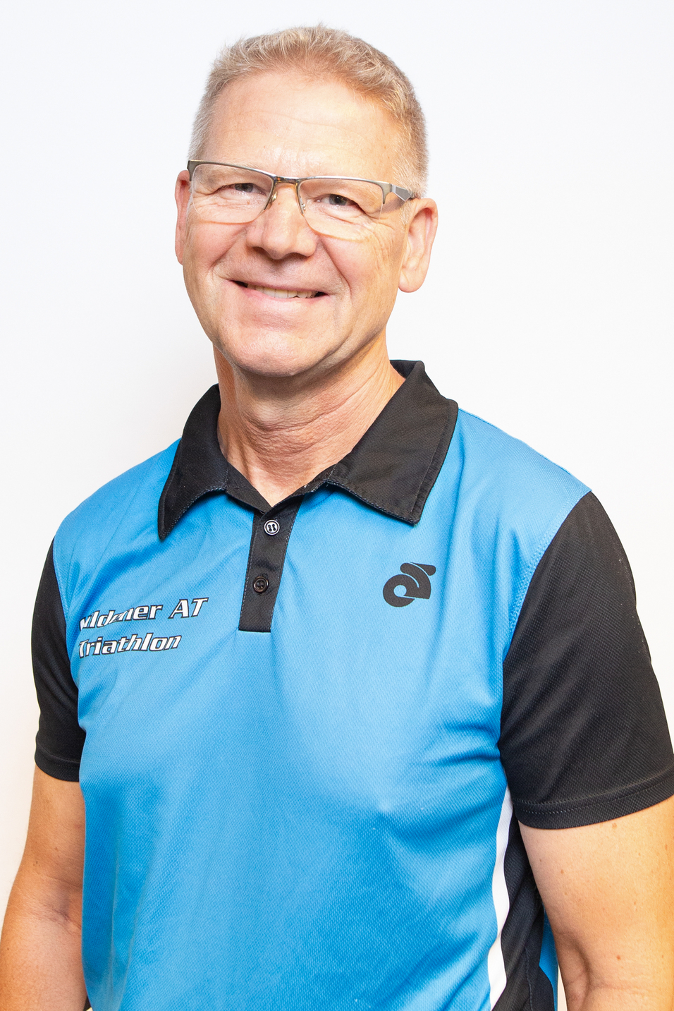 Portrait eines Mannes mit Brille, der ein blau-schwarzes Sportshirt tr&auml;gt, l&auml;chelt vor einem neutralen Hintergrund.