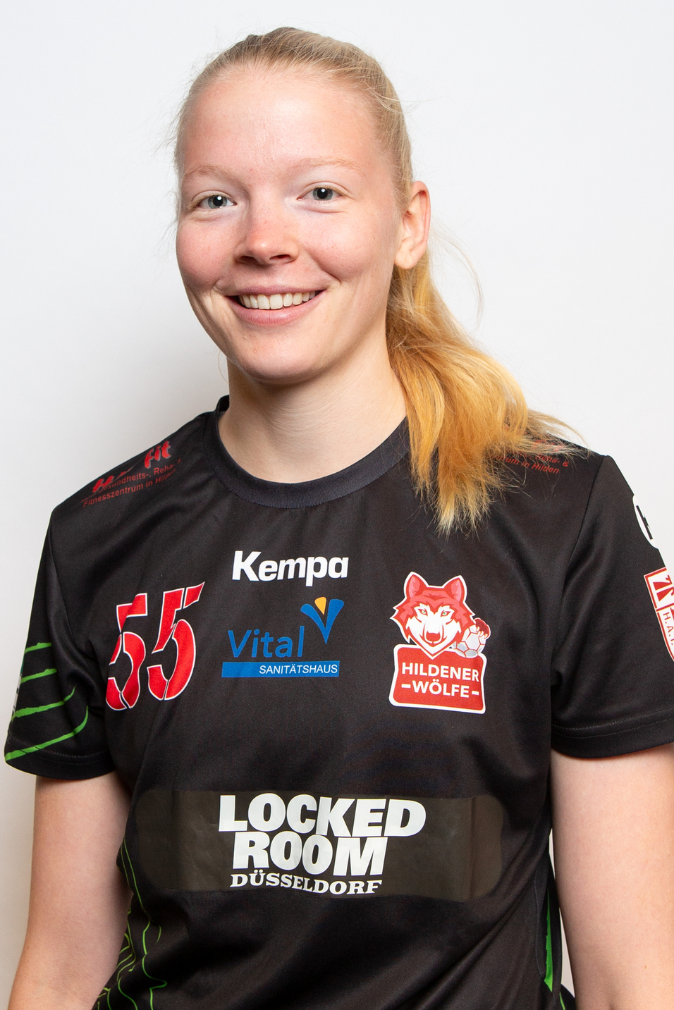 L&auml;chelnde Sportlerin in schwarzem Trikot mit Logos, pr&auml;sentiert sich vor einem neutralen Hintergrund.