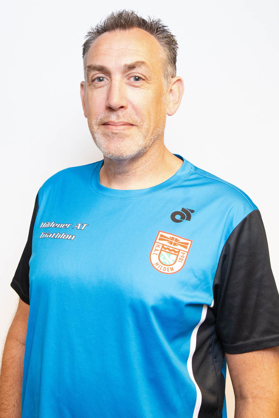 Portr&auml;t eines Mannes mit kurzen Haaren, der ein blau-schwarzes Sporttrikot tr&auml;gt, vor schlichter heller Wand.