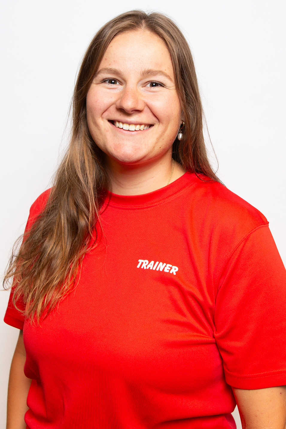 L&auml;chelnde Frau mit langen Haaren tr&auml;gt ein rotes T-Shirt mit dem Aufdruck &bdquo;TRAINER&ldquo;. Hintergrund ist wei&szlig;.