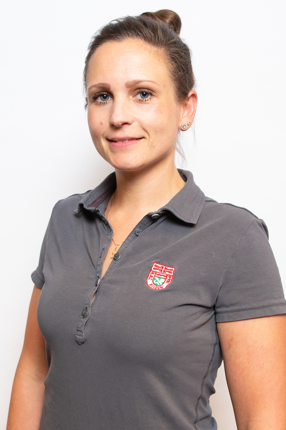 Portr&auml;t einer Frau mit kurzen Haaren, die ein graues Poloshirt mit emblembeschm&uuml;ckt tr&auml;gt. Hintergrund ist neutral und hell.