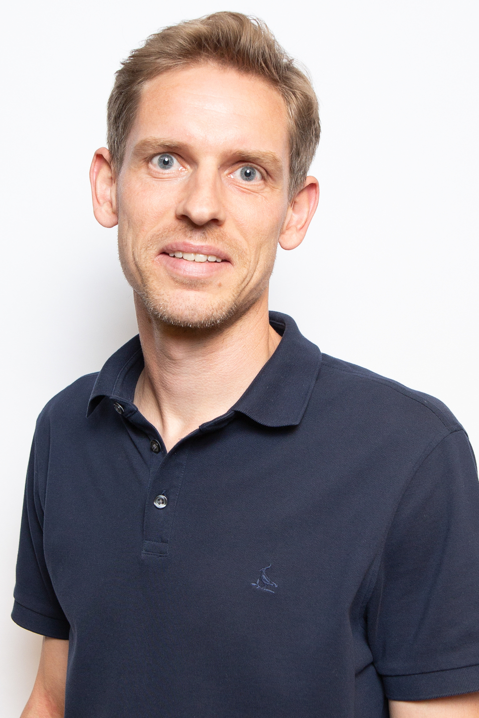 Portr&auml;t eines Mannes mit kurzem, blondem Haar, der ein dunkles Polo-Shirt tr&auml;gt und in die Kamera l&auml;chelt.