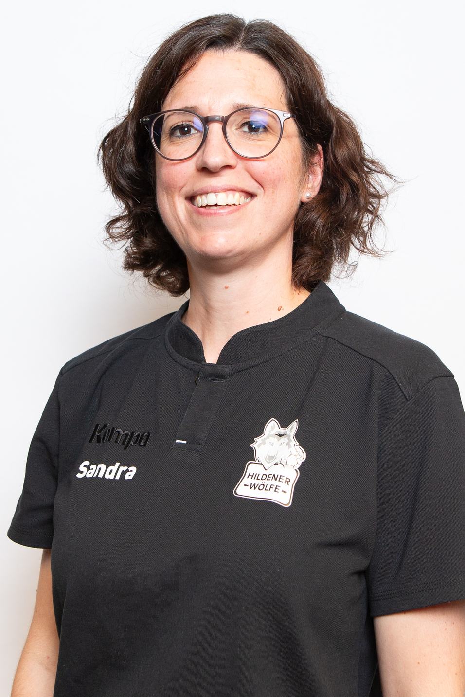 Portr&auml;t einer l&auml;chelnden Frau mit lockigen Haaren in einem schwarzen Poloshirt mit dem Schriftzug &bdquo;Hildener W&ouml;lfe&ldquo;.