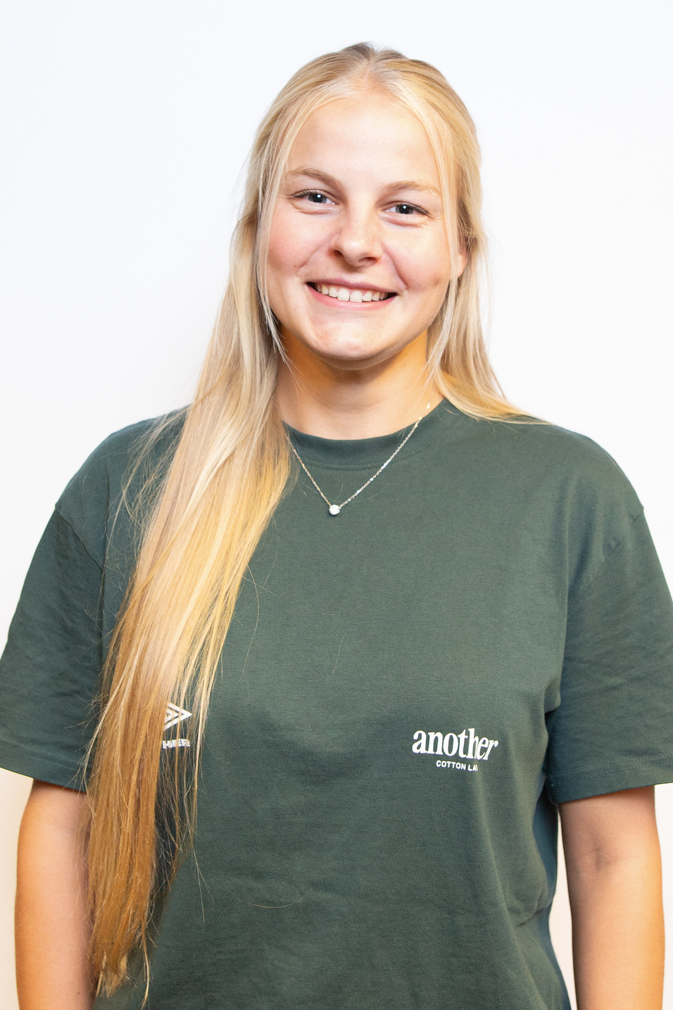 L&auml;chelnde junge Frau mit langen blonden Haaren tr&auml;gt ein gr&uuml;nes T-Shirt mit Aufschrift und steht vor einem wei&szlig;en Hintergrund.