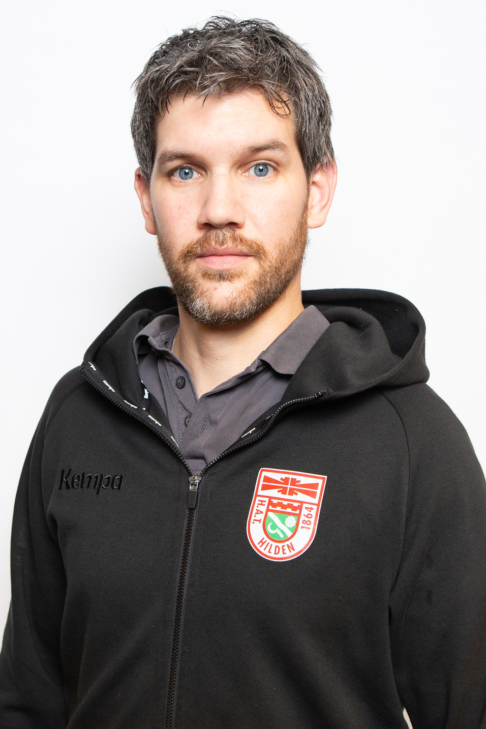 Portr&auml;t eines Mannes mit kurzem, grauem Haar und Bart, tr&auml;gt einen schwarzen Kapuzenpullover mit Logo. Hintergrund ist wei&szlig;.