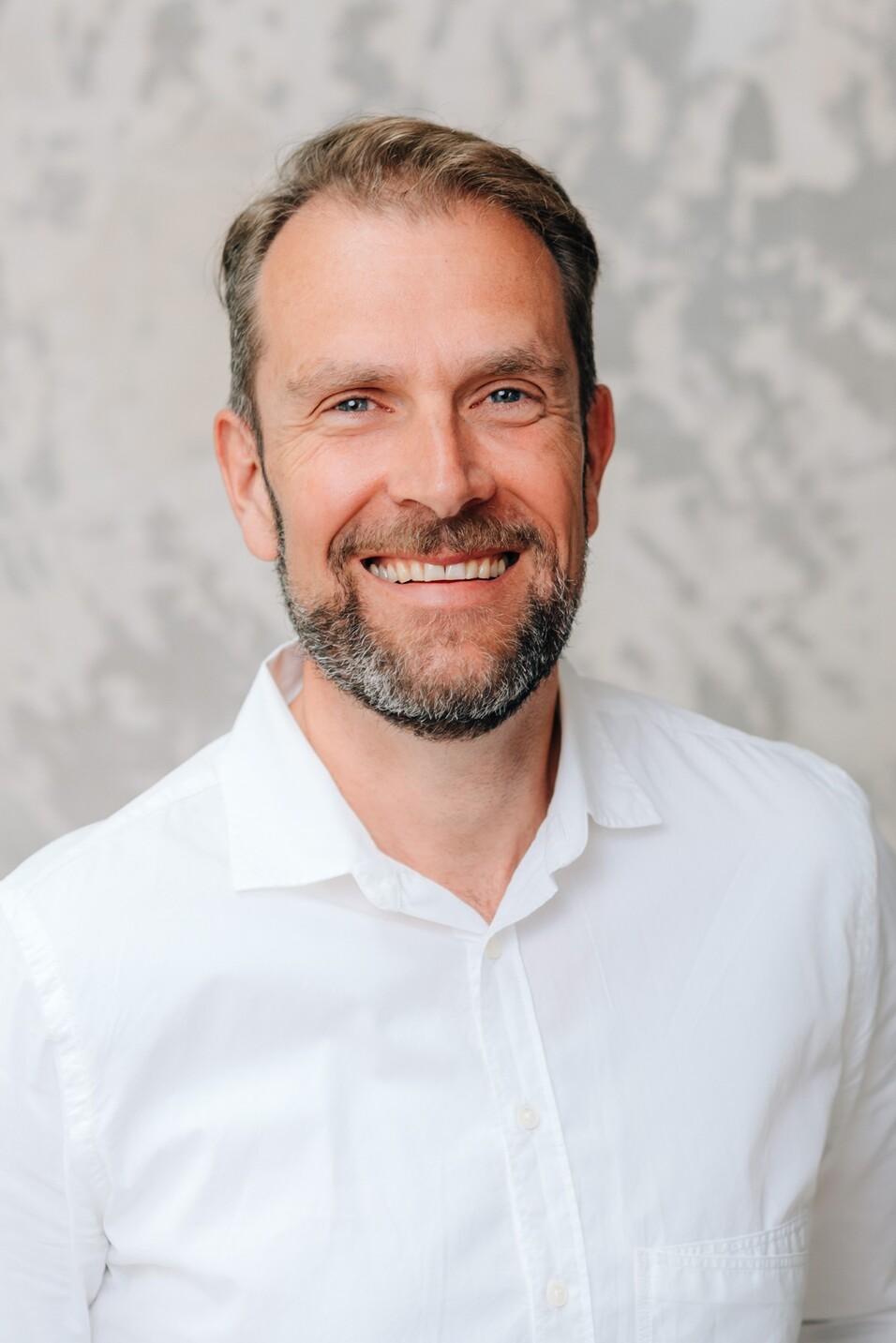 Lächelnder Mann mit Bart in weißem Hemd vor einer neutralen, strukturierten Hintergrundwand.