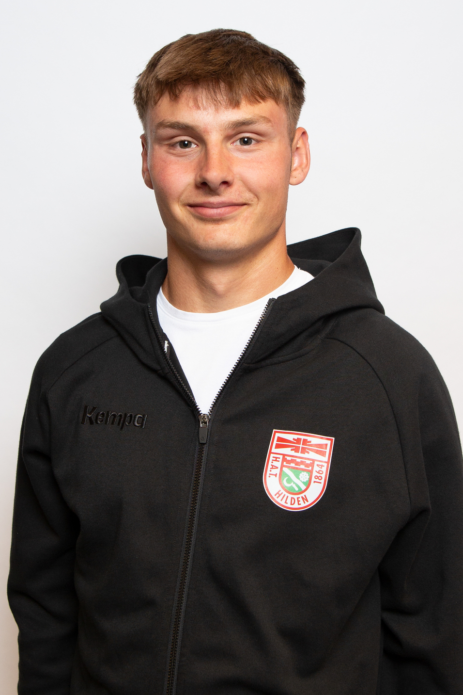 Portr&auml;t eines jungen Mannes in einem schwarzen Kapuzenpulli mit Logo, l&auml;chelnd vor einem wei&szlig;en Hintergrund.