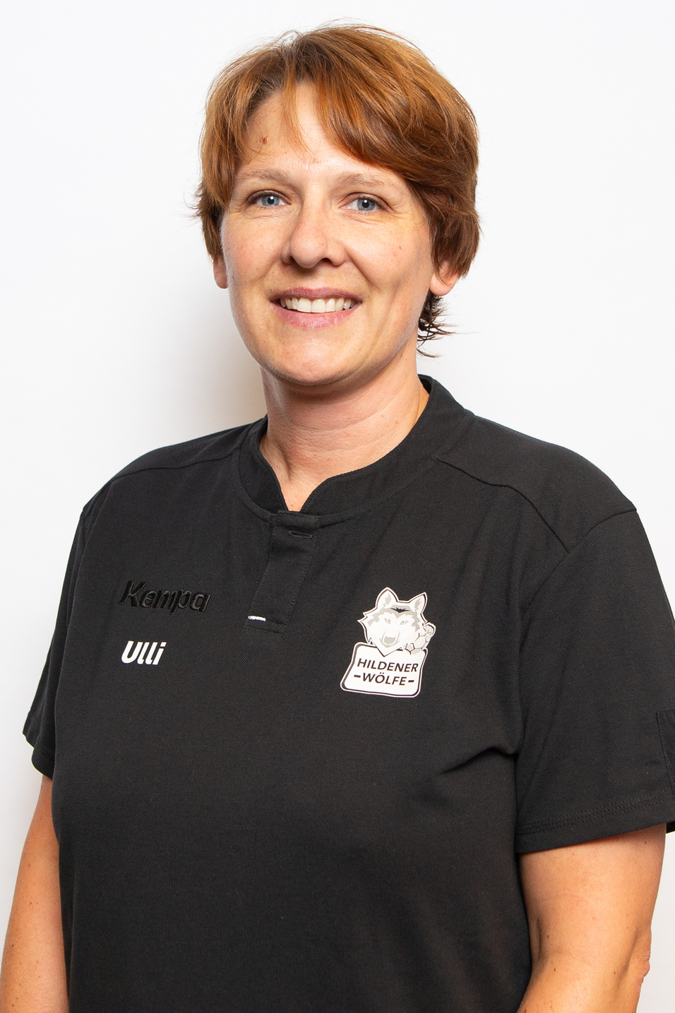 Portr&auml;t einer l&auml;chelnden Person in einem schwarzen Shirt mit Aufdruck "Hildener W&ouml;lfe", vor hellem Hintergrund.