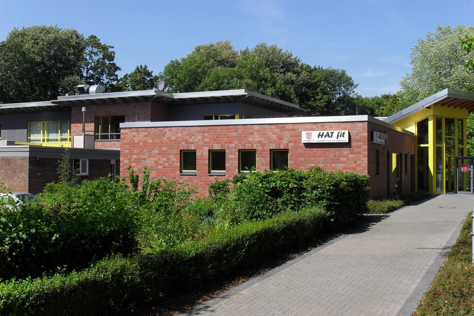 Modernes Geb&auml;ude mit Backsteinfassade und gr&uuml;nen Pflanzen, beschattet von B&auml;umen, unter klarem Himmel.
