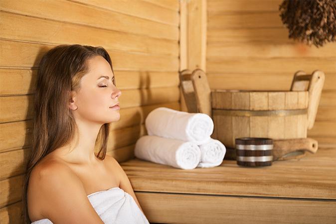 Frau entspannt in einer Sauna, umgeben von einem Holzbottich, frischen Handtüchern und einer ruhigen Atmosphäre.