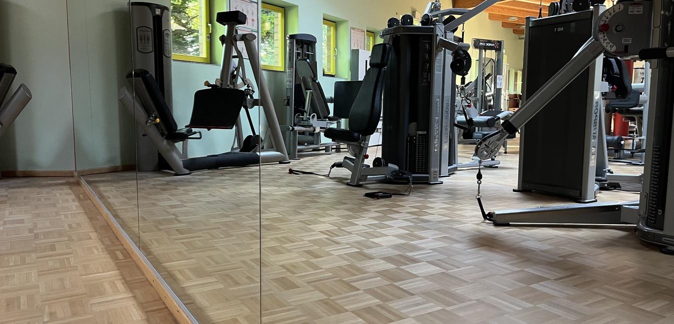 Reflexion im Spiegel eines Fitnessraums mit verschiedenen Trainingsgeräten und Holzfußboden.