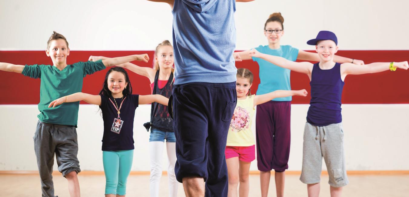 Eine Gruppe von Kindern und einem Lehrer zeigt Tanzbewegungen in einem Trainingsraum mit Holzfußboden.