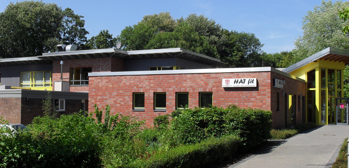 Modernes Geb&auml;ude mit Backsteinfassade und gr&uuml;nen Pflanzen, beschattet von B&auml;umen, unter klarem Himmel.