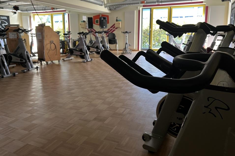 Blick auf einen Fitnessraum mit mehreren Spinning-Bikes und Holzfußboden, ausgestattet mit großen Fenstern.